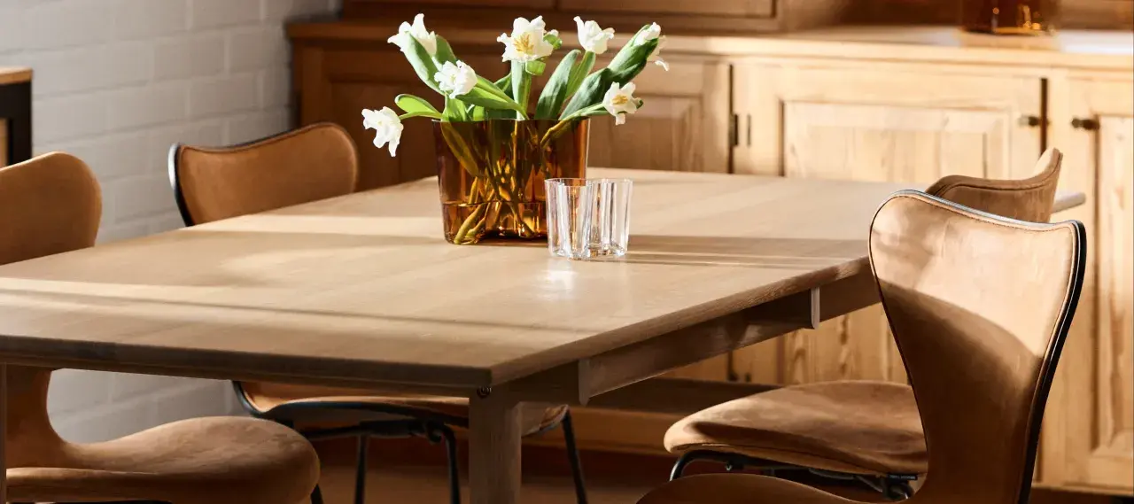 Ett ljust träbord med klassiska designstolar i läder. En vas med vita blommor och glas står på bordet.