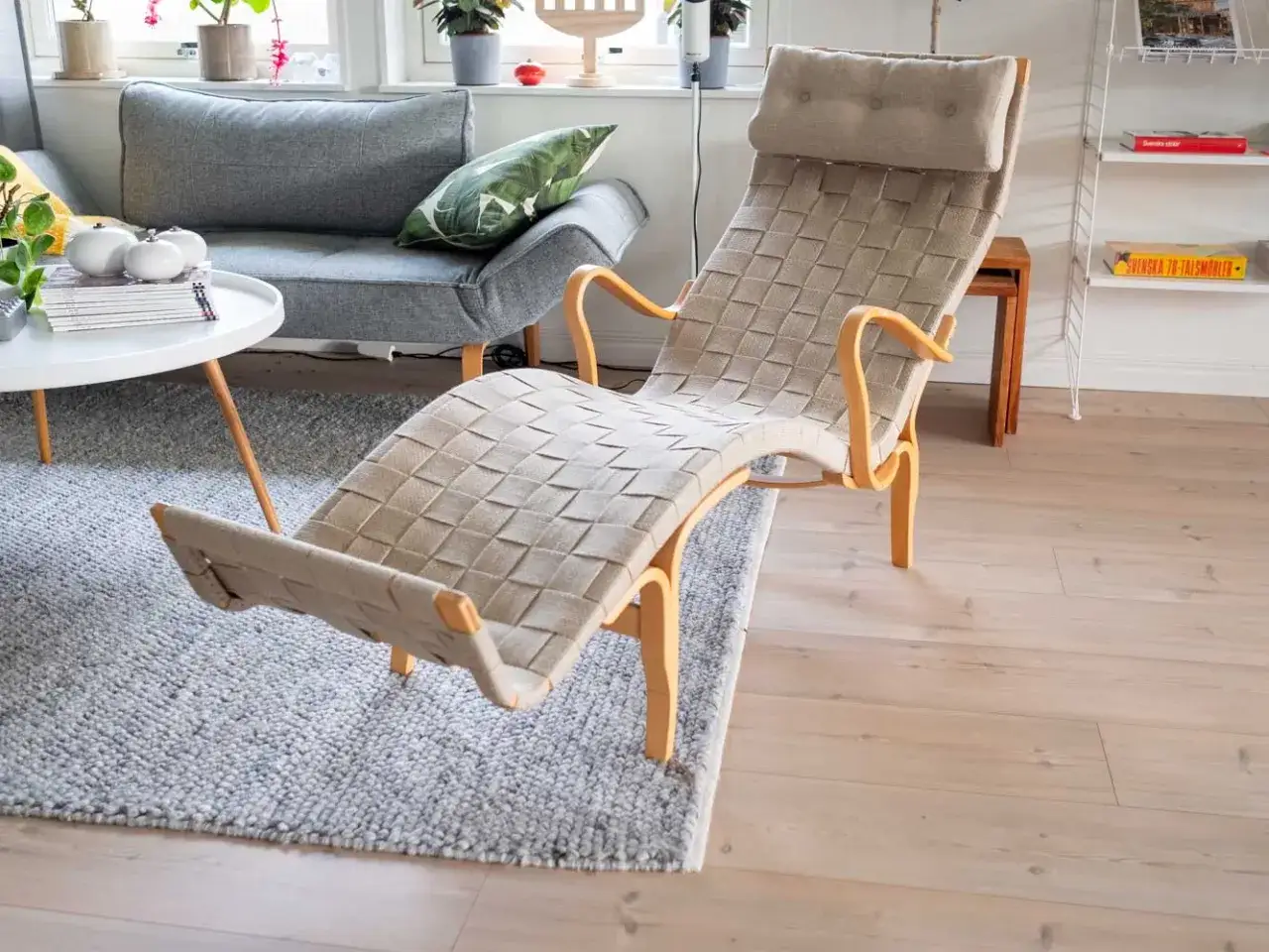 En klassisk bruno mathsson liggfåtölj i beige flätat tyg med böjda träarmstöd, placerad på en grå matta i ett ljust vardagsrum.