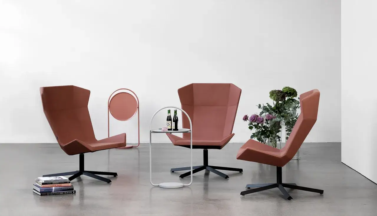 Tre moderna, terrakottaf&auml;rgade f&aring;t&ouml;ljer fr&aring;n Johanson Design st&aring;r i ett rum med en rund rosa piedestal, ett litet sidobord med flaskor och en blomsterbukett.